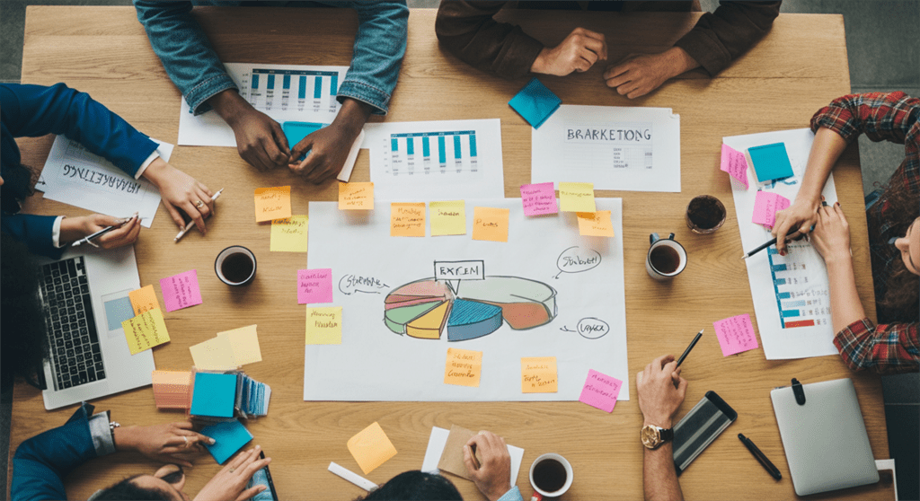 Uma imagem fotográfica de profissionais criativos discutindo ideias em volta de uma grande mesa, cheia de gráficos coloridos, post-its e dispositivos digitais, simbolizando um avanço na estratégia de marketing.